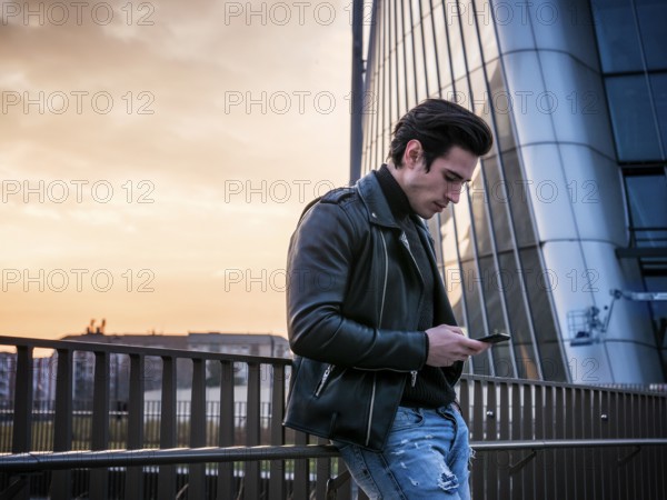 One handsome young man in urban setting in modern city, standing, wearing black leather jacket and