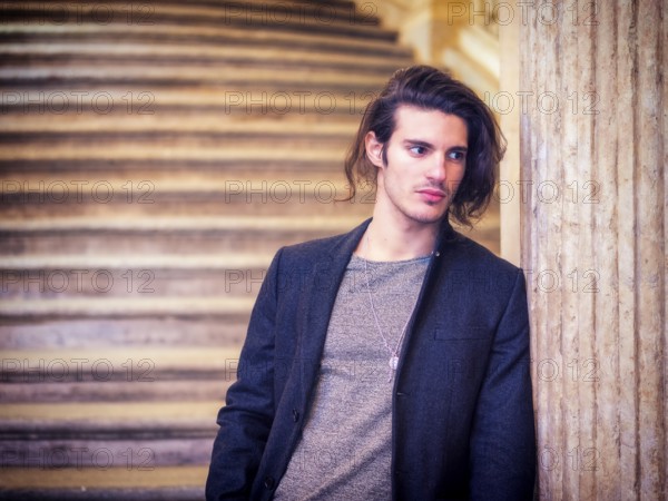 A young, long-haired attractive man standing next to a marble column, leaning onto it, wearing