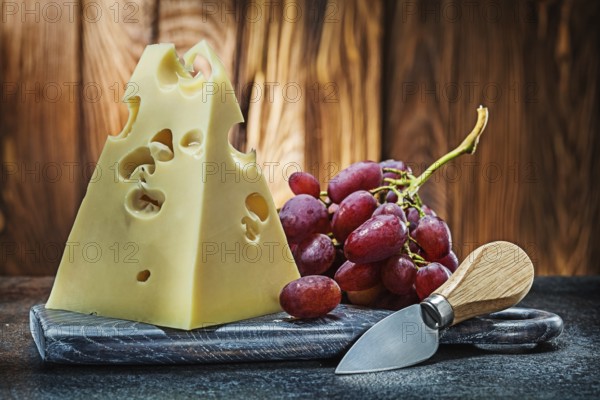Piece of maasdam cheese on vintage carving board knife and bunch of rose grape old wooden