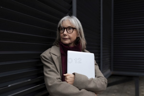 A middle-aged businesswoman with glasses is standing against a dark wall, holding a laptop. She