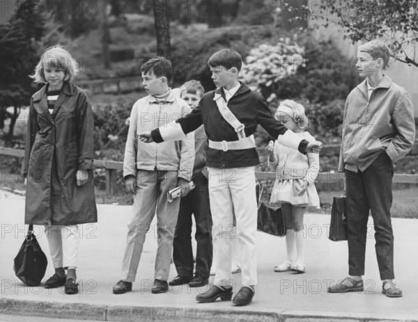 Junior safety patrol in the 1960s. A teenage boy stands with his arms out and signals to the