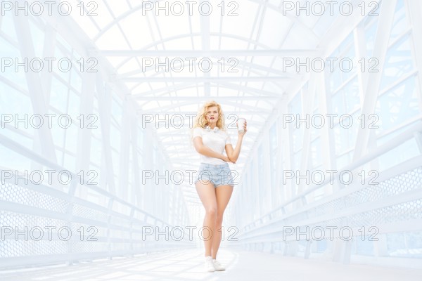 Beautiful young woman with music headphones standing on the bridge with a takeaway coffee mug and