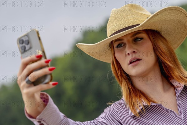 A beautiful redhead model uses her cellphone while posing in a country setting wearing cowgirl