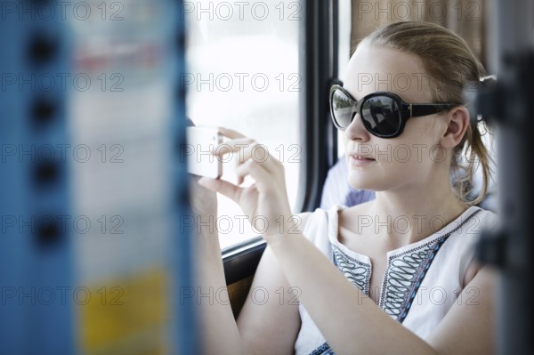 Attractive young woman wearing sunglasses holding up and photographing at her mobile on a bus or