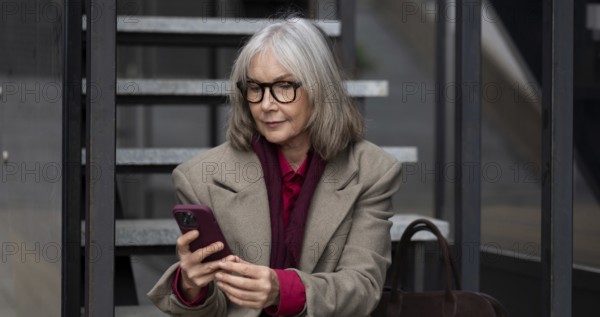 A middle-aged businesswoman dressed in a stylish coat and glasses is engaged with her smartphone
