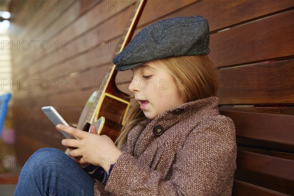 Blond kid girl playing smartphone winter beret and guitar on wooden background