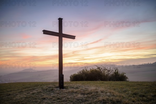 A cross stands in front of a colourful sunset in a peaceful landscape, Bisberg, Watterdingen,