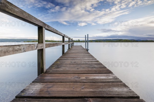 Long wooden footbridge leads into a quiet lake under a partly cloudy sky, Naturfreundehaus,