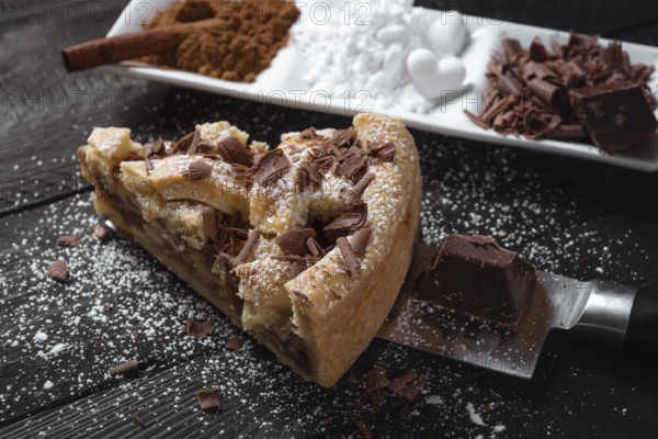 Slice of apple pie with powdered sugar and chocolate, on a knife, on a black rustic table and a