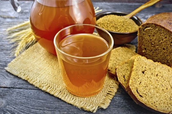 Kvass in glassful and glass jug on burlap, malt in a bowl, rye bread and ears on a wooden plank