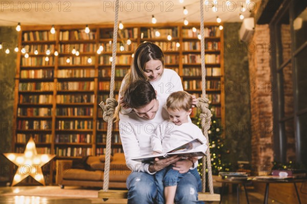 Family with baby relaxing at home swing seat in cosy dark living room on Christmas eve. Family