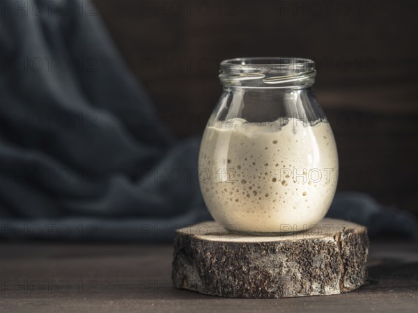 Active wheat sourdough starter in glass jar on brown wooden background. Starter for sourdough bread
