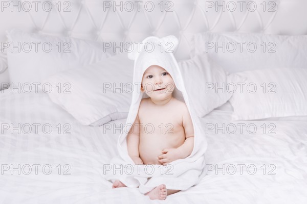 Adorable newborn baby boy with brown eyes sitting wrapped in a white towel with ears in light bed.