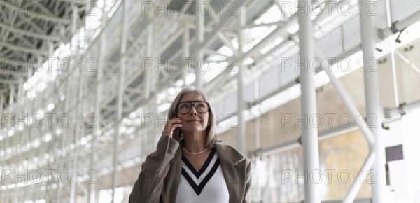 A middle-aged businesswoman with glasses is engaged in a phone conversation while strolling through
