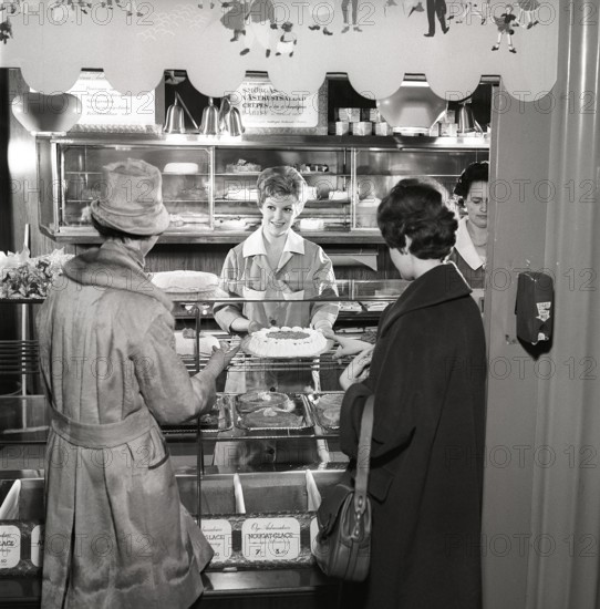 1960s coffee shop. Two women are standing in front of the counter that is full with freshly baked
