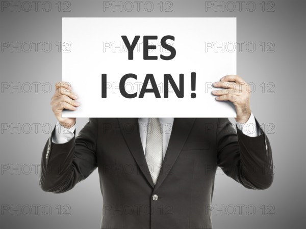 A business man holding a paper in front of his face with the message yes i can