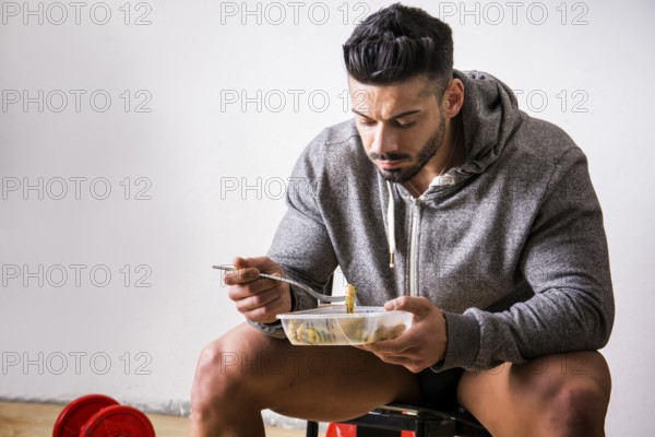 Hungry muscular young man gulping down food looking at plastic container without pausing as he