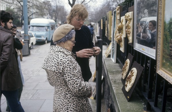 London 1982. A view from a London street with an elderly lady looking at art hanged on the fence to
