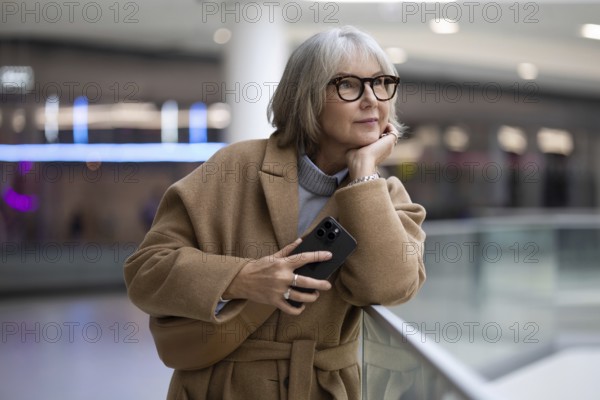 A woman with stylish glasses leans against a railing, thoughtfully holding her smartphone. She