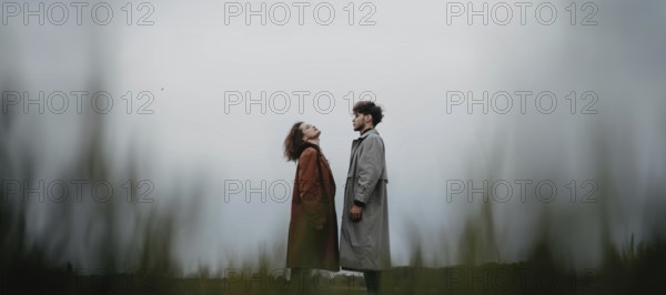 A couple standing in a field, each looking upwards, dressed in long coats with a sense of