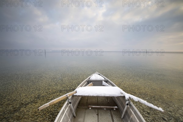 A snow-covered boat on a calm lake with pebbles on the shore under a cloudy sky, Strandbad Hörnle,