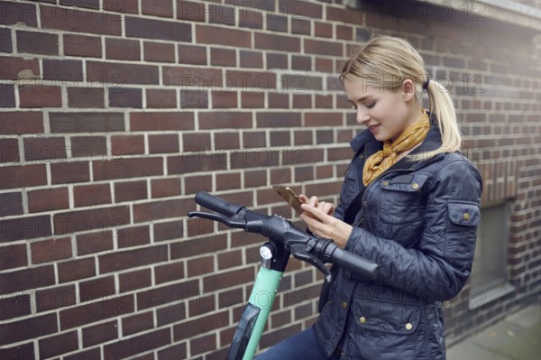 Trendy young blond woman in leather jacket standing at her e-scooter booking a ride, ecological