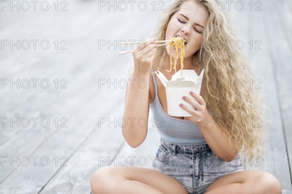 Happy young woman with long curly hair holding a lunch box and eating noodles from a Chinese snack