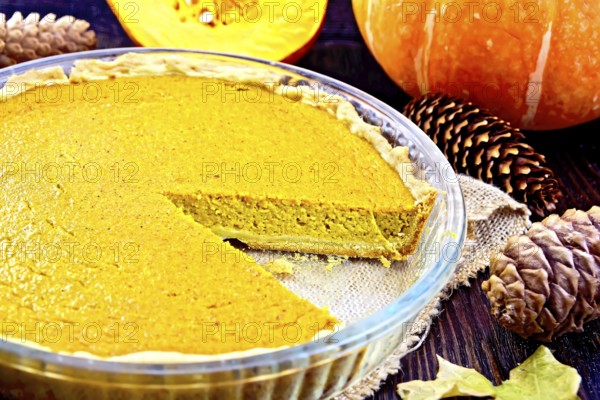 Pumpkin pie in a glass pan on sackcloth, pumpkins, pine cones and maple leaf on the background of