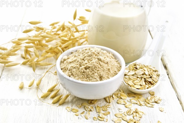 Flour oat in bowl, milk in a jug, oatmeal in spoon, oaten stalks on the background of light wooden