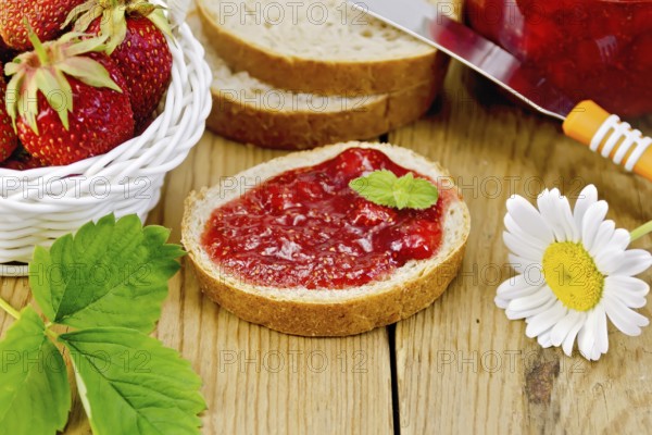 Bread with strawberry jam, a jar of jam, knife, strawberries in a wicker basket, chamomile on a
