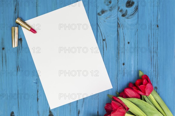 Red tulips, lipstick and a white sheet of paper for your greetings on the background of blue wooden