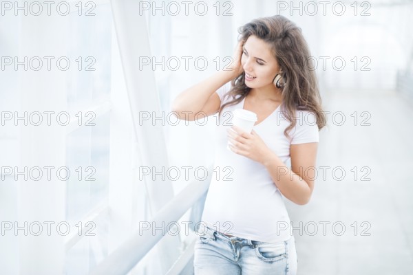 Happy young woman with music headphones standing on the bridge with a takeaway coffee mug, looking