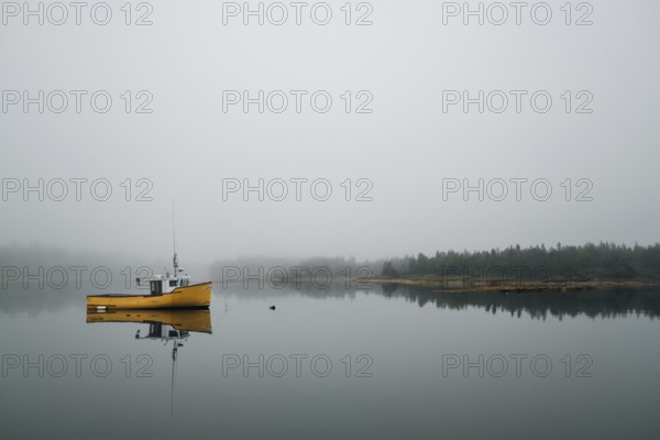 A commercial fishing boat is reflected in the still calm water of a protected bay as light fog