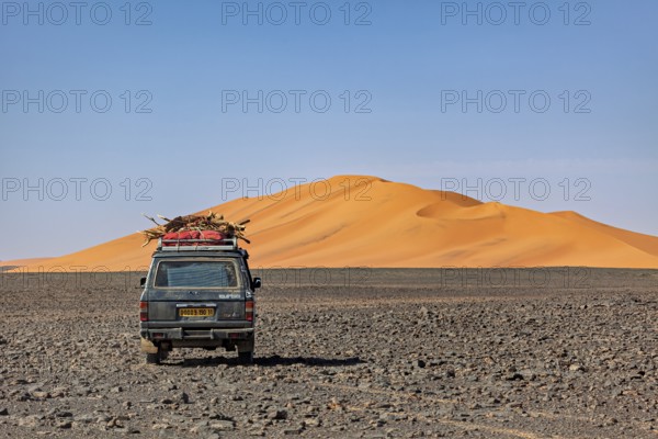 Off-road vehicle leads through a rocky plain in front of large sand dunes under a blue sky, By