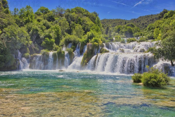 Waterfalls flow into an emerald green river, surrounded by dense forest and a bright blue sky, Krka