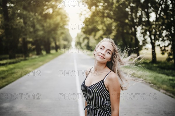 Woman standing on a road surrounded by trees, sunlight filtering through, creating a relaxed summer
