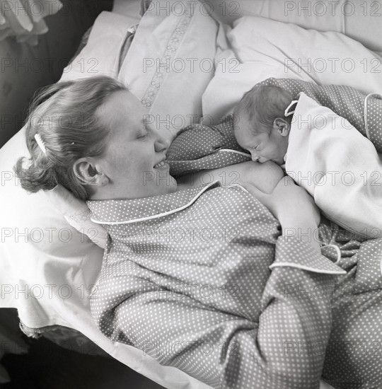 1950s mother nursing her baby. A young woman breastfeeding her baby while lying in bed at home.