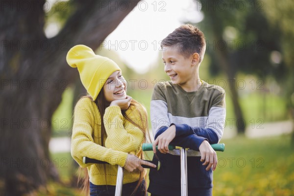 Twins 10 years old in bright clothes spend time actively together in autumn park riding kick