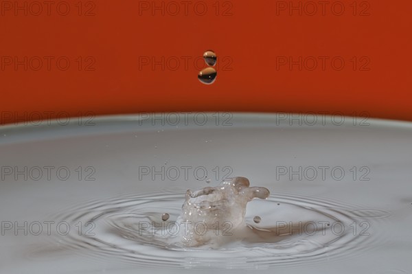 A beige water droplet hits the water surface and creates a small splash on an orange background,