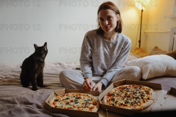 Woman sitting on the bed with pizza boxes and a black cat in a nighttime cozy setting