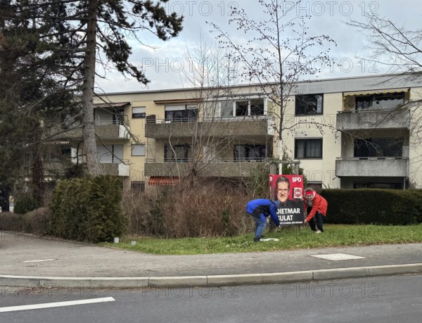 A scene in a residential area with trees, pavement and people working on an election poster,