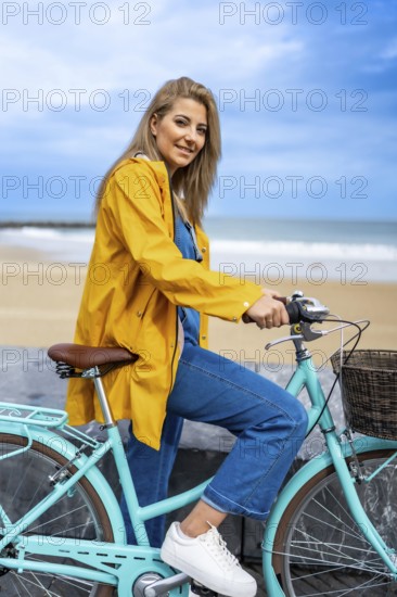 Young woman with yellow raincoat sitting on a turquoise vintage bicycle by the beach, enjoying a
