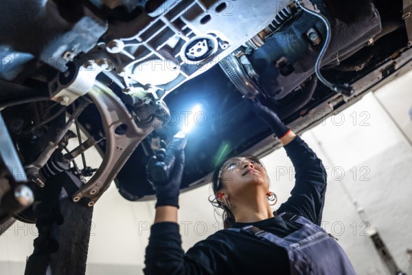 Female auto mechanic inspecting the underside of a car in a repair shop, using a flashlight to