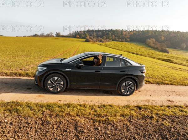 A driver sits in a black car on a dirt track, surrounded by green meadows, electric car VW ID5,
