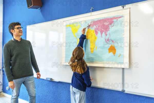 Smiling teacher watching elementary school student pointing at a world map during geography class