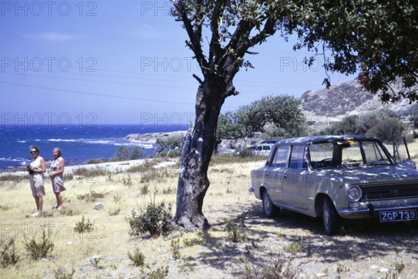 Two women standing by coast with Fiat 124 car parked in shade of olive tree, Mediterranean Sea,