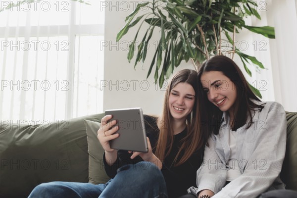 Two teenager sister girls are sitting at green sofa in living room and watching some funny video at