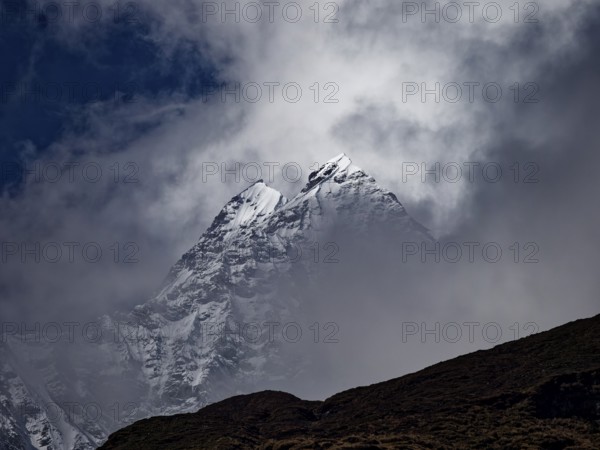 Two brightly lit snowy peaks peeking out of the clouds behind a dark ridge, Annapurna region,