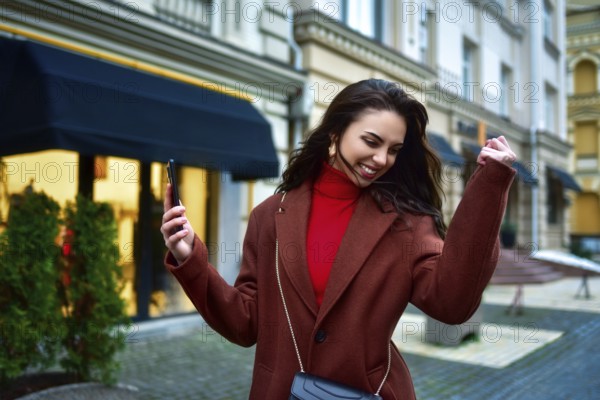 Beautiful Overjoyed young brunette woman with long hair in a red coat celebrating good news holding
