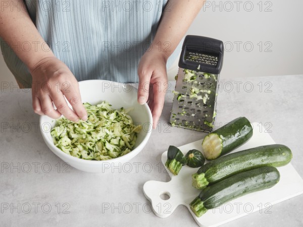 Woman with her hands in a bowl of grated zucchini, other fresh zucchini on wooden board and